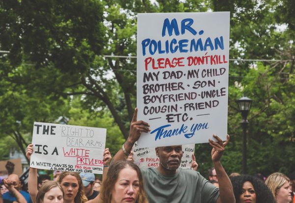 Image shows people holding protest signs. One sign says "Mr. policeman please don't kill my day, my child, brother, uncle, cousin, friend, etc. thank you." Another says, " the right to bear arms is a white privilege." 