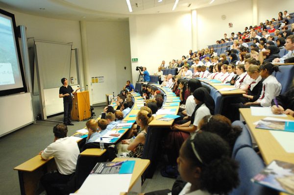 Photo shows a lecture hall with many students sitting in rows, facing the front of the room where a professor stands near a podium. 