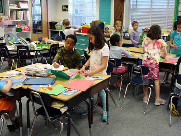 Photo of elementary school students standing by their desks working with construction paper. 