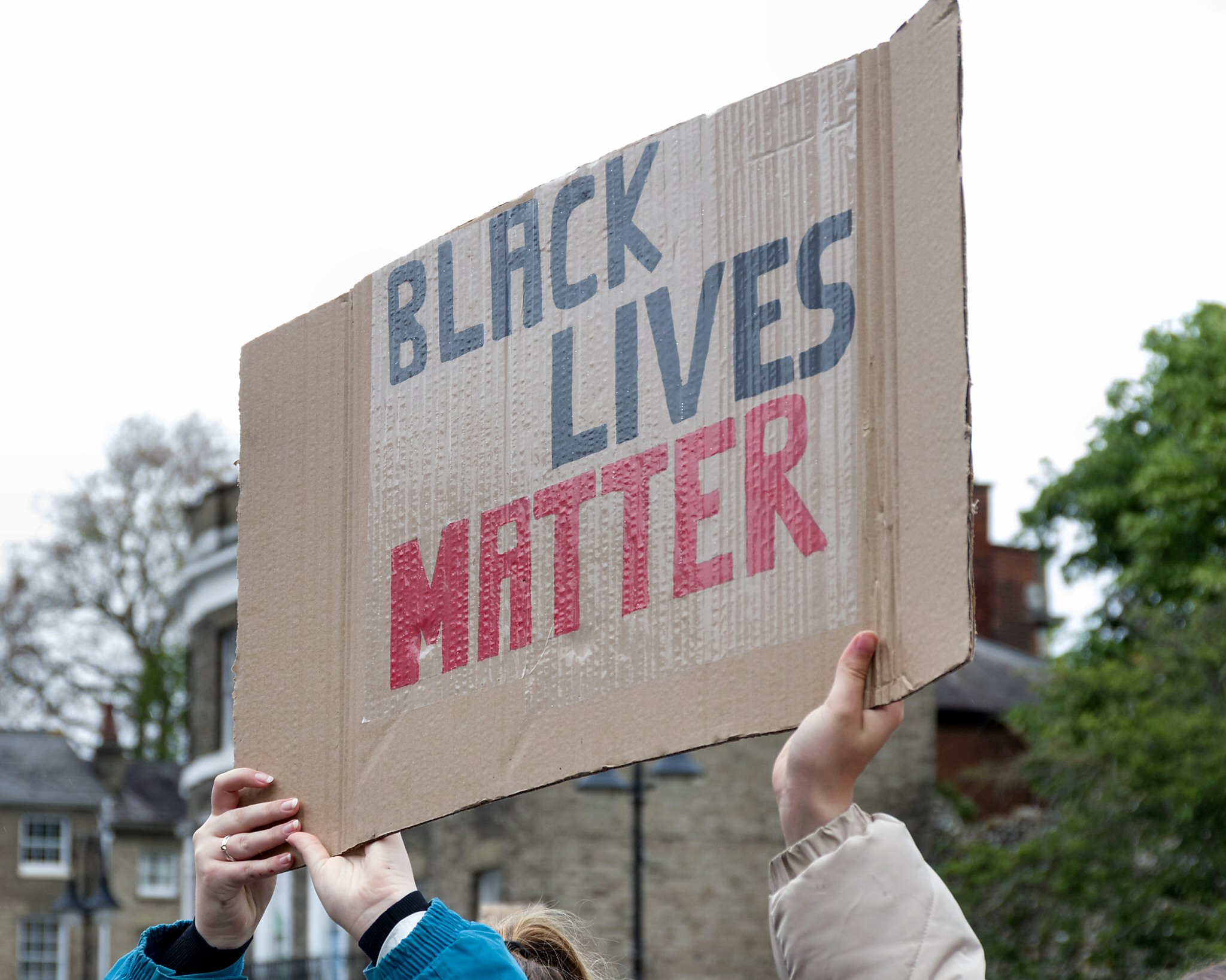 Black Lives Matter is written on a cardboard sign and held up in the air by a pair of white hands.