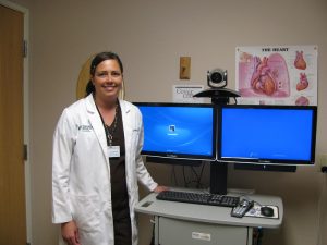 Doctor stands next to telehealth set up: two large monitors and a camera