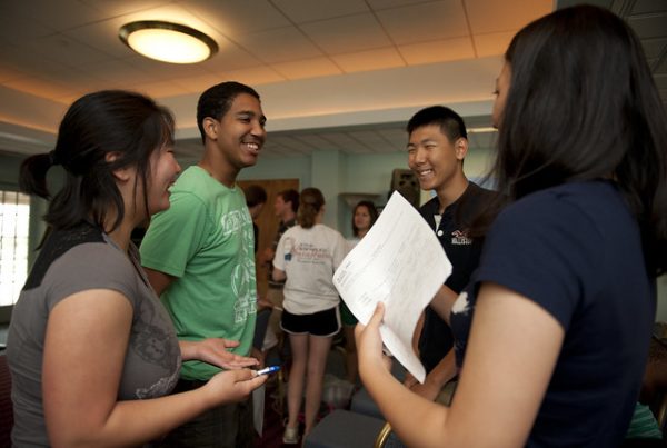 Four young adults stand in a circle facing each other. They are all smiling. One is holding a piece of paper. 