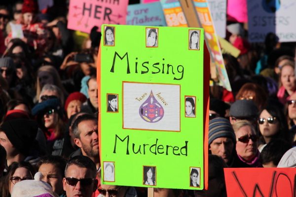 Photo shows a crowd of people holding signs. The sign in focus is green and says "Missing Murdered" and shows photos of Indigenous women. 
